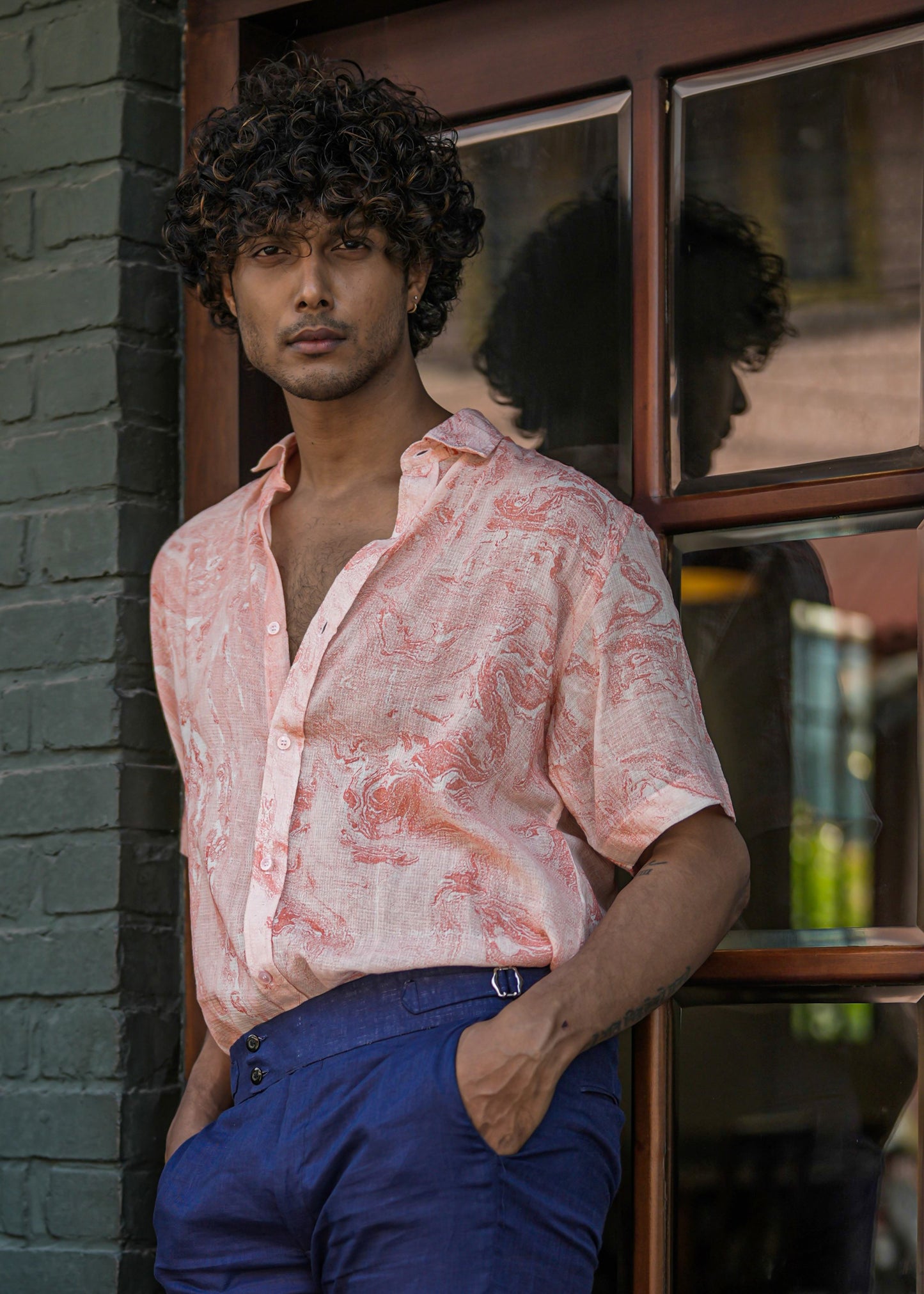 Male model standing confidently in front of a charming dark brown wood cafe door, set against a military green brick wall backdrop. He wears a stylish Dobby cotton marble printed half-sleeve shirt in vibrant orange from the brand Mod Maven, paired impeccably with navy blue formal fit trousers. The ensemble exudes a perfect blend of casual elegance, making it a versatile and trendy fashion statement.