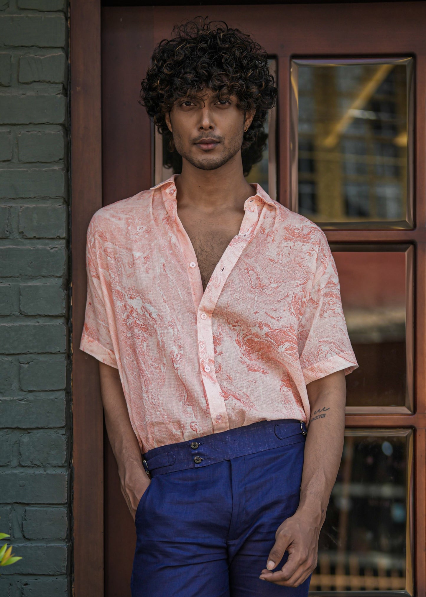 Male model standing confidently in front of a charming dark brown wood cafe door, set against a military green brick wall backdrop. He wears a stylish Dobby cotton marble printed half-sleeve shirt in vibrant orange from the brand Mod Maven, paired impeccably with navy blue formal fit trousers. The ensemble exudes a perfect blend of casual elegance, making it a versatile and trendy fashion statement.