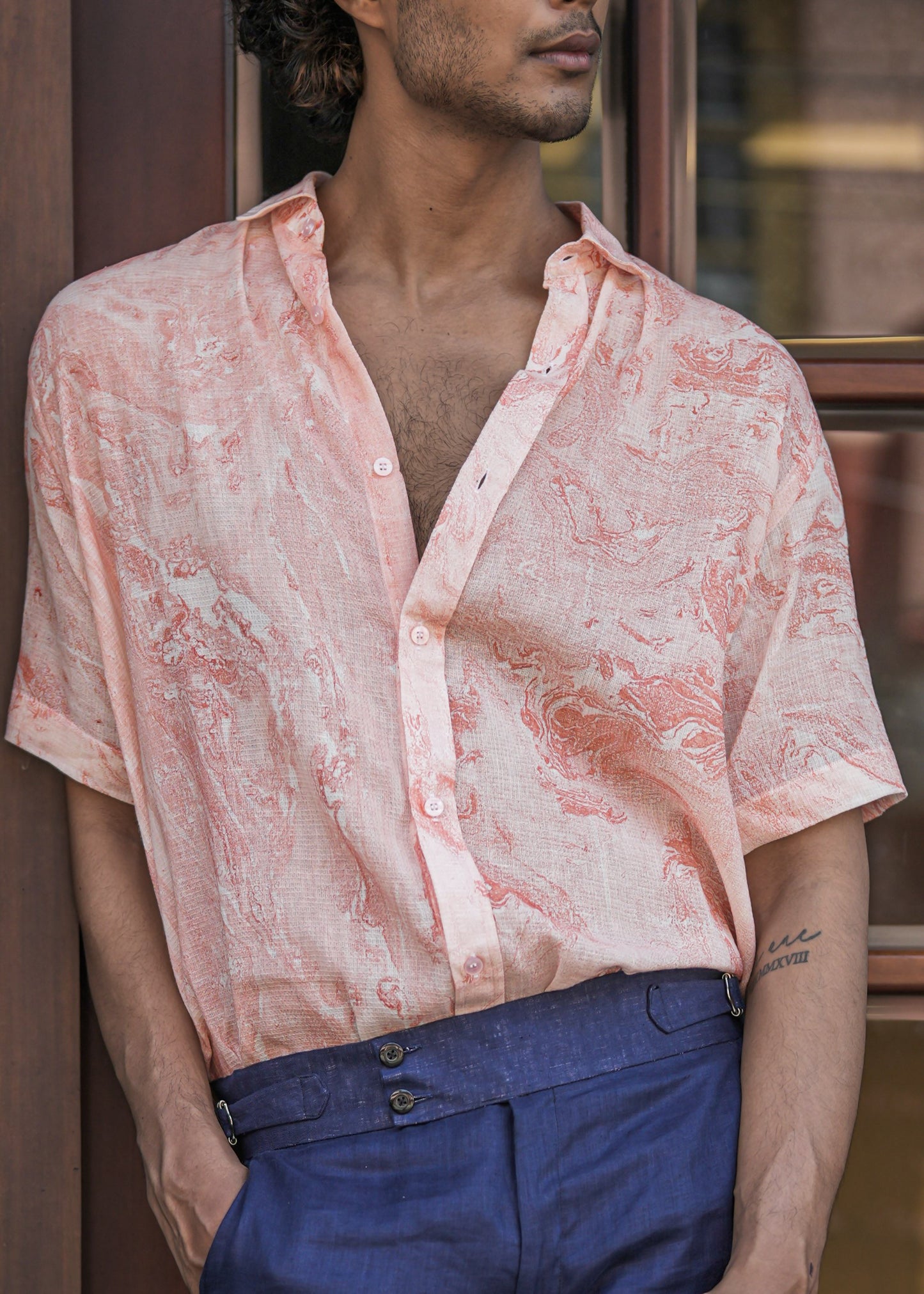 Close-up shot of a male model standing confidently in front of a dark brown wood cafe door, set against a military green brick wall. The model is wearing a stylish Dobby cotton marble printed half-sleeve shirt in a vibrant orange color from the renowned brand Mod Maven. The shirt's intricate texture and captivating pattern are highlighted in the image, showcasing its unique design. Paired impeccably with navy blue formal fit trousers, the ensemble exudes a perfect blend of modern fashion and sophistication.