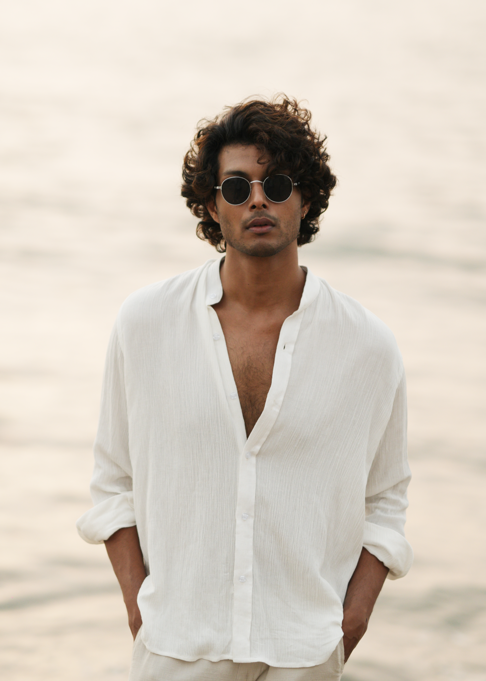 Mod Maven's white crushed cotton full sleeve shirt exudes casual elegance, paired with off-white trousers for a comfortable fit. The model stands against a serene beach backdrop, wearing stylish shades, showcasing the brand's effortlessly chic and laid-back aesthetic.