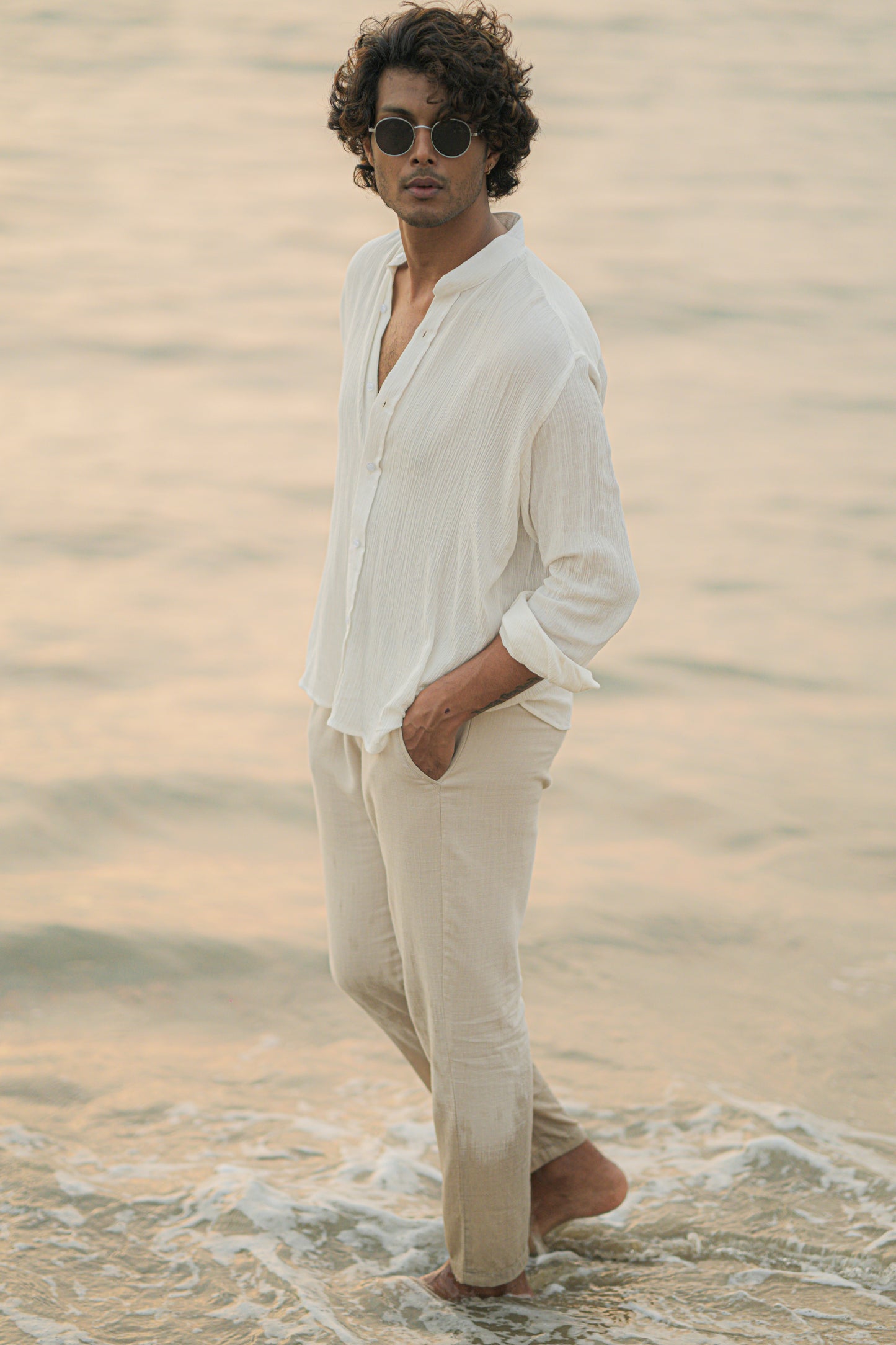 Mod Maven's white crushed cotton full sleeve shirt exudes casual elegance, paired with off-white trousers for a comfortable fit. The model stands against a serene beach backdrop, wearing stylish shades, showcasing the brand's effortlessly chic and laid-back aesthetic.