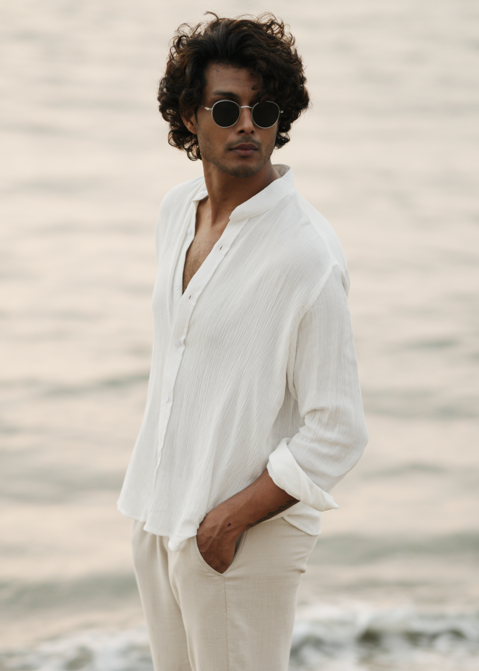 Mod Maven's white crushed cotton full sleeve shirt exudes casual elegance, paired with off-white trousers for a comfortable fit. The model stands against a serene beach backdrop, wearing stylish shades, showcasing the brand's effortlessly chic and laid-back aesthetic.
