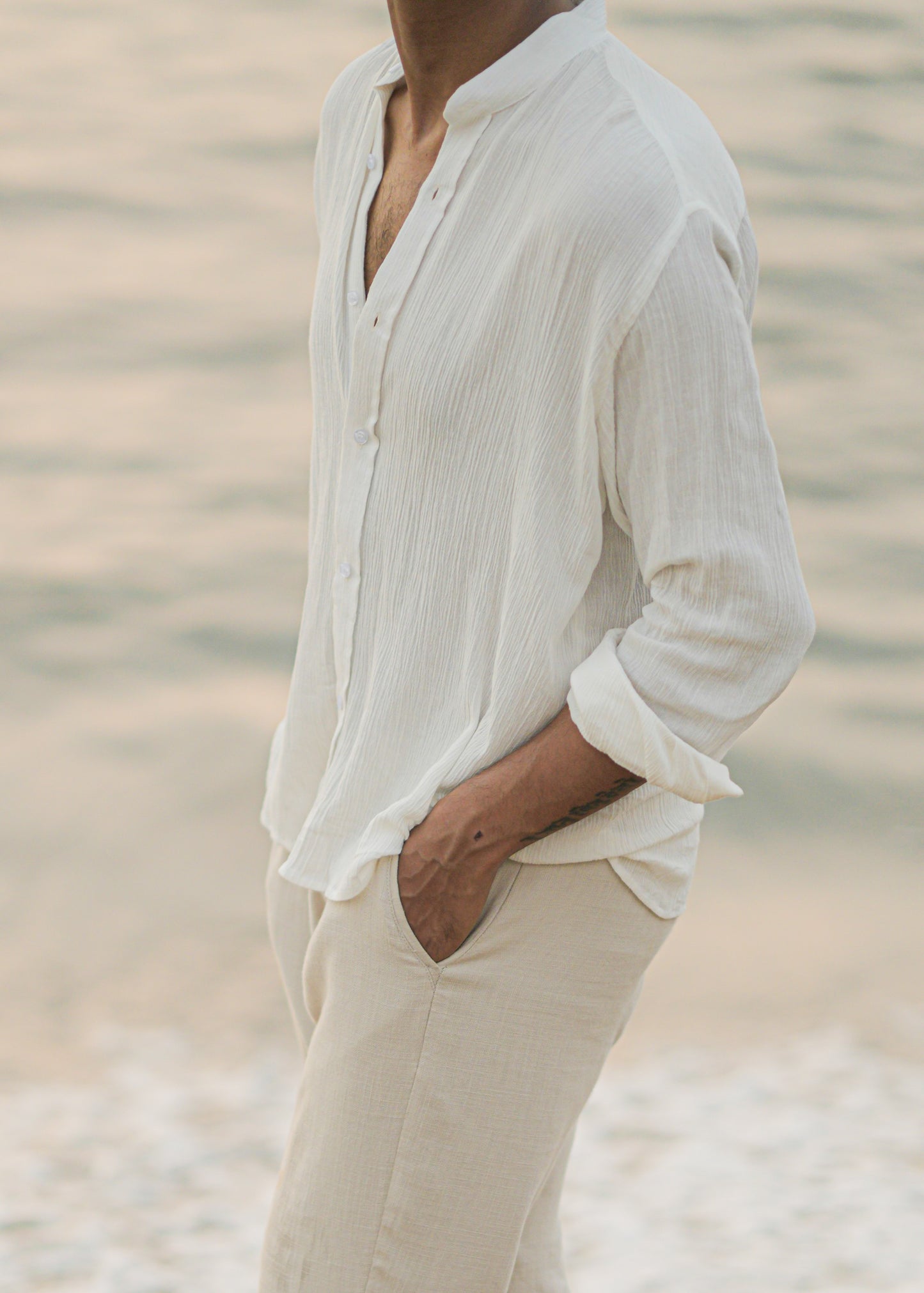 Mod Maven's white crushed cotton full sleeve shirt exudes casual elegance, paired with off-white trousers for a comfortable fit. Image shows a close up shot of model in the crushed cotton shirt, standing against a serene beach backdrop, showcasing the brand's effortlessly chic and laid-back aesthetic.