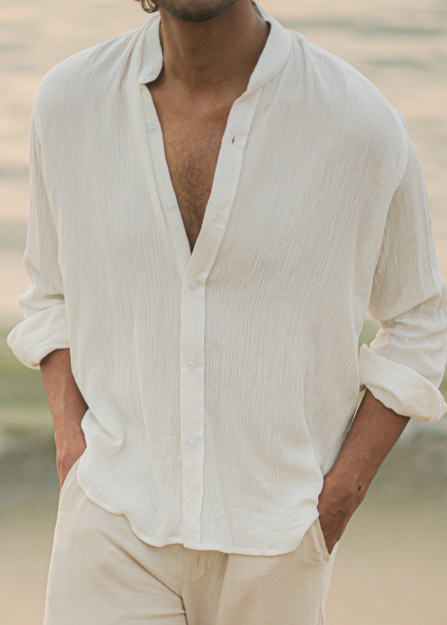 Mod Maven's white crushed cotton full sleeve shirt exudes casual elegance, paired with off-white trousers for a comfortable fit. Image shows a close up shot of model in the crushed cotton shirt, standing against a serene beach backdrop, showcasing the brand's effortlessly chic and laid-back aesthetic.