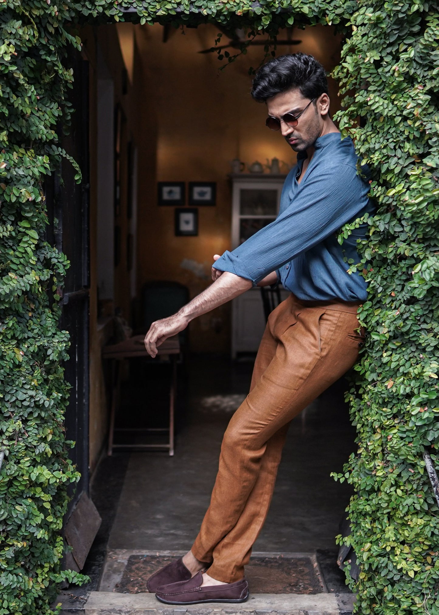 Mod Maven Prussian Blue Crushed Cotton Full Sleeve Shirt paired with Mud Brown formal trousers. Model standing at a coffee shop entrance, adjusting his sleeves, wearing shades, showcasing a semi formal look.
