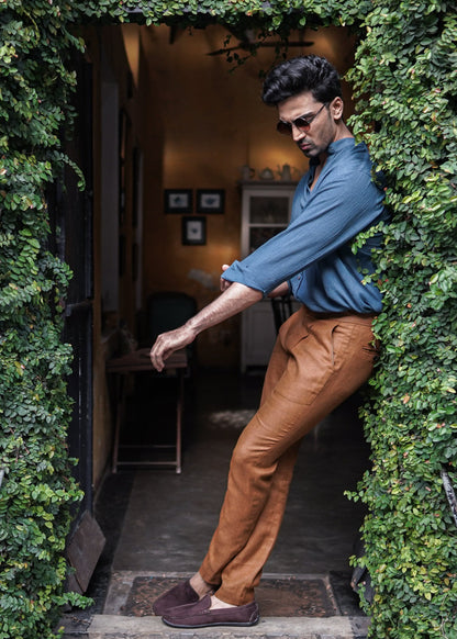 Mod Maven Prussian Blue Crushed Cotton Full Sleeve Shirt paired with Mud Brown formal trousers. Model standing at a coffee shop entrance, adjusting his sleeves, wearing shades, showcasing a semi formal look.