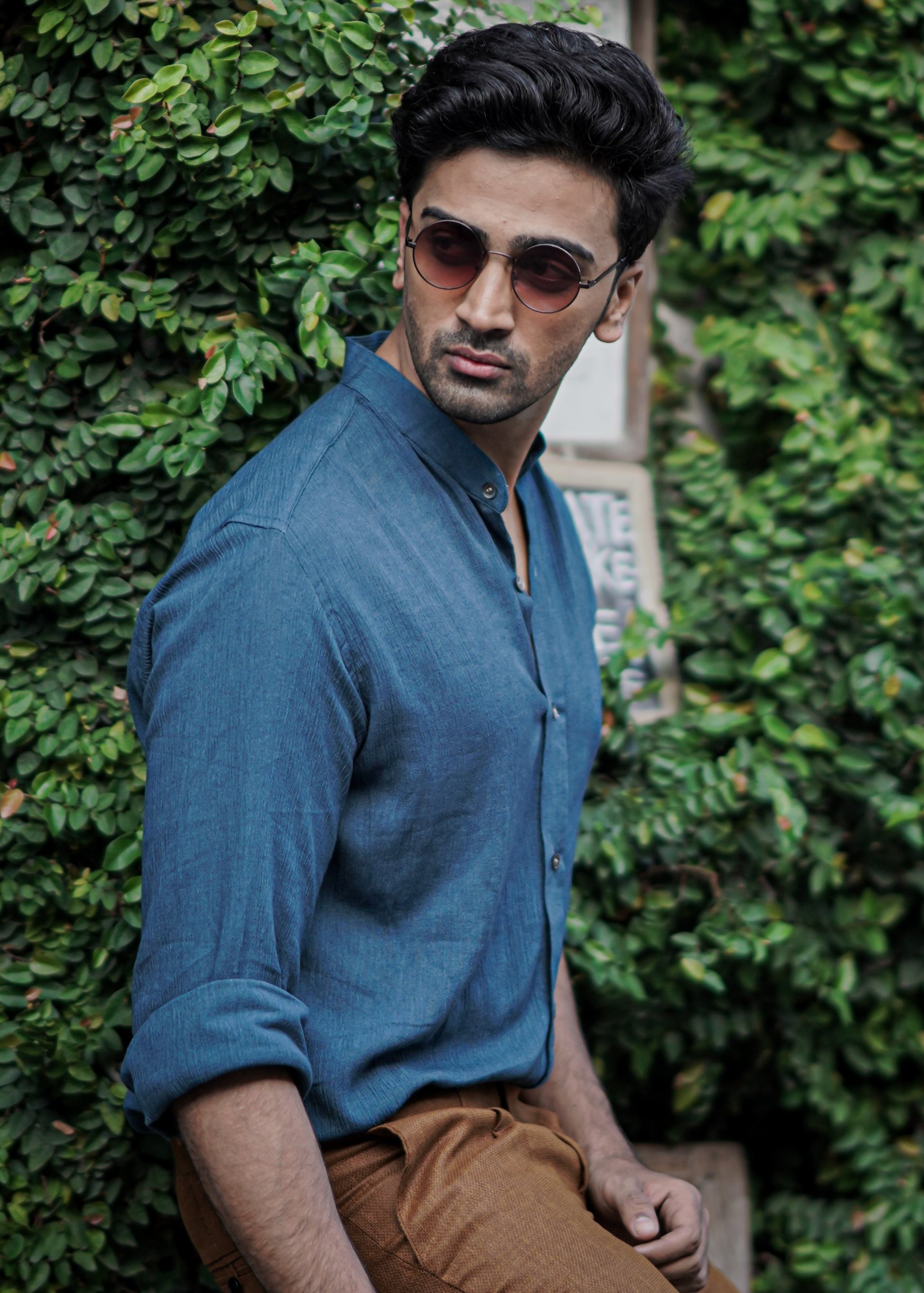Male model stands against a backdrop of lush greenery wearing sunglasses, a full-sleeve crushed cotton shirt in Prussian blue by Mod Maven, paired with mud brown formal trousers, exuding urban style amidst a street setting.