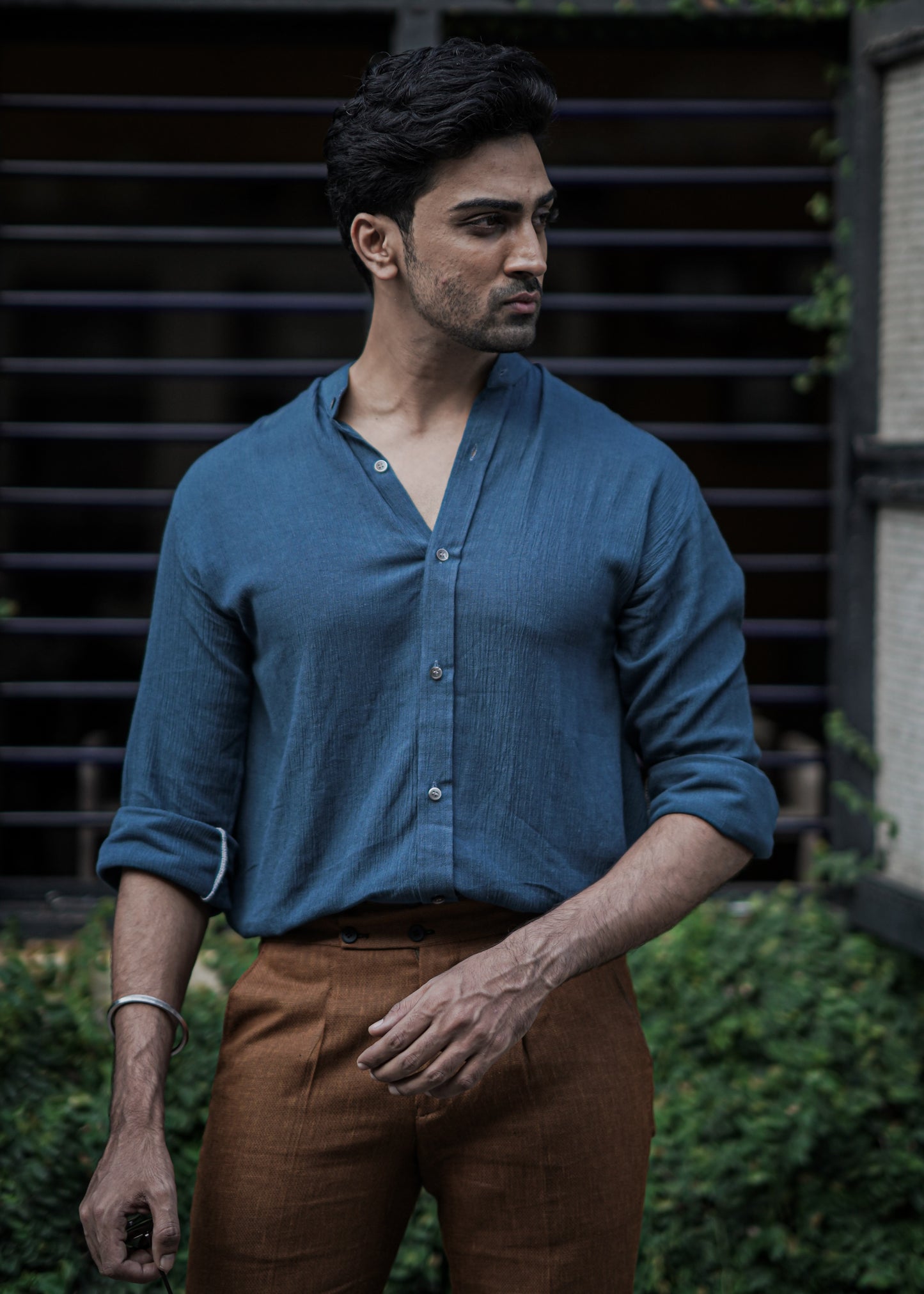 Male model stands against a backdrop of lush greenery, a full-sleeve crushed cotton shirt in Prussian blue by Mod Maven, paired with mud brown formal trousers, exuding urban style amidst a street setting.