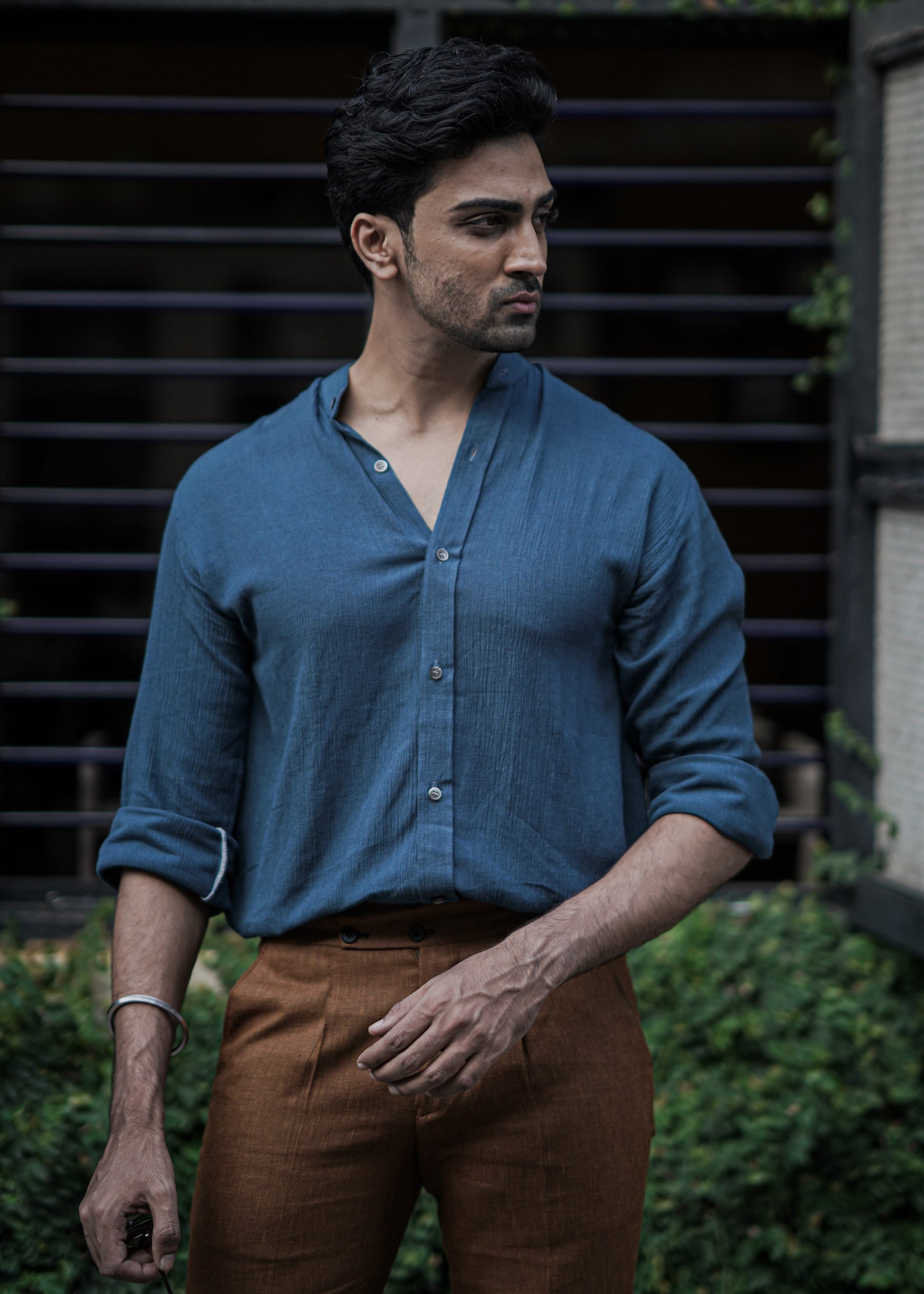 Male model stands against a backdrop of lush greenery, a full-sleeve crushed cotton shirt in Prussian blue by Mod Maven, paired with mud brown formal trousers, exuding urban style amidst a street setting.