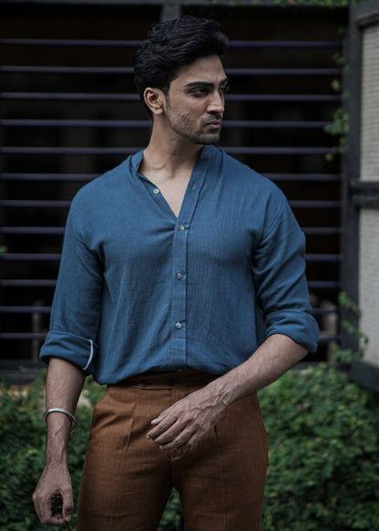 Male model stands against a backdrop of lush greenery, a full-sleeve crushed cotton shirt in Prussian blue by Mod Maven, paired with mud brown formal trousers, exuding urban style amidst a street setting.