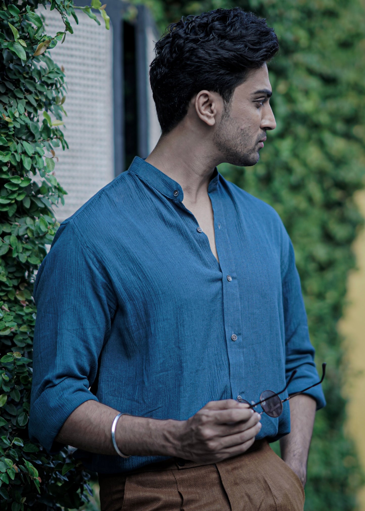 Male model stands against a backdrop of lush greenery with sunglasses in hand, a full-sleeve crushed cotton shirt in Prussian blue by Mod Maven, paired with mud brown formal trousers, exuding urban style amidst a street setting.