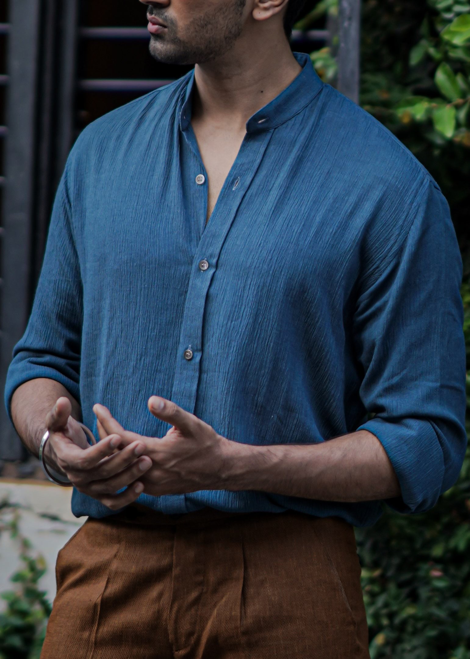 Close up image of Male model standing against a backdrop of lush greenery, focused on the texture of full-sleeve crushed cotton shirt in Prussian blue by Mod Maven, paired with mud brown formal trousers, exuding urban style amidst a street setting.