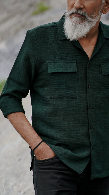 A distinguished older man with a gray beard and stylishly combed gray hair is wearing an army green, double-pocket, full-sleeve shirt from Mod Maven. He pairs the shirt with dark trousers and accessorizes with a sleek black wristwatch, standing confidently against a textured, natural background. The picture focuses on the collar and pocket detail