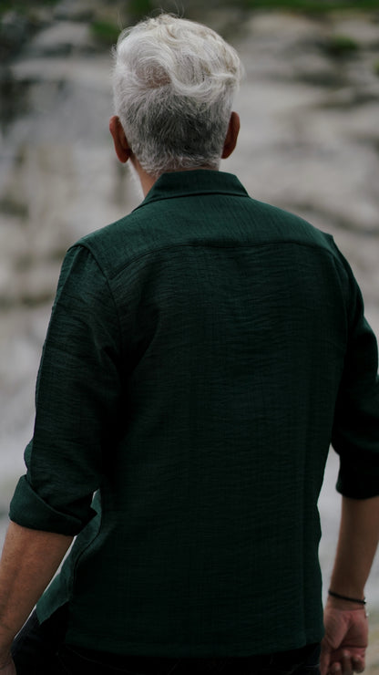 Back view of a stylish older man with a neatly combed gray hair is showcasing an army green, double-pocket, full-sleeve shirt from Mod Maven. Set against a rocky backdrop conveying a sense of adventure and elegance. His outfit is completed with black trousers, black shoes,  emphasizing his effortlessly cool and sophisticated style.