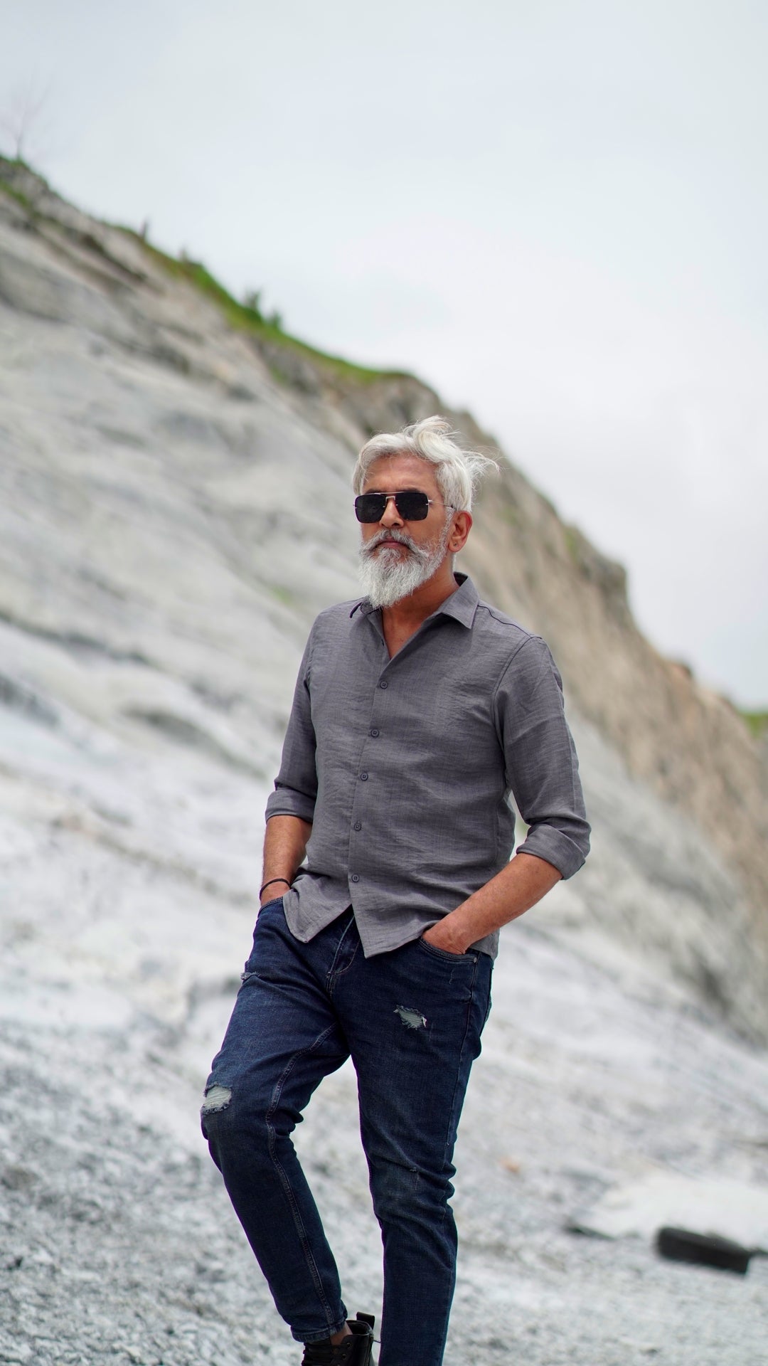 A distinguished older male model with a gray beard and stylishly combed gray hair is wearing a charcoal grey, full-sleeve shirt from Mod Maven. He pairs the shirt with dark trousers and accessorizes with black sunglasses and a sleek black wristwatch, standing confidently against a textured, natural background. The overall look exudes modern sophistication and timeless style.