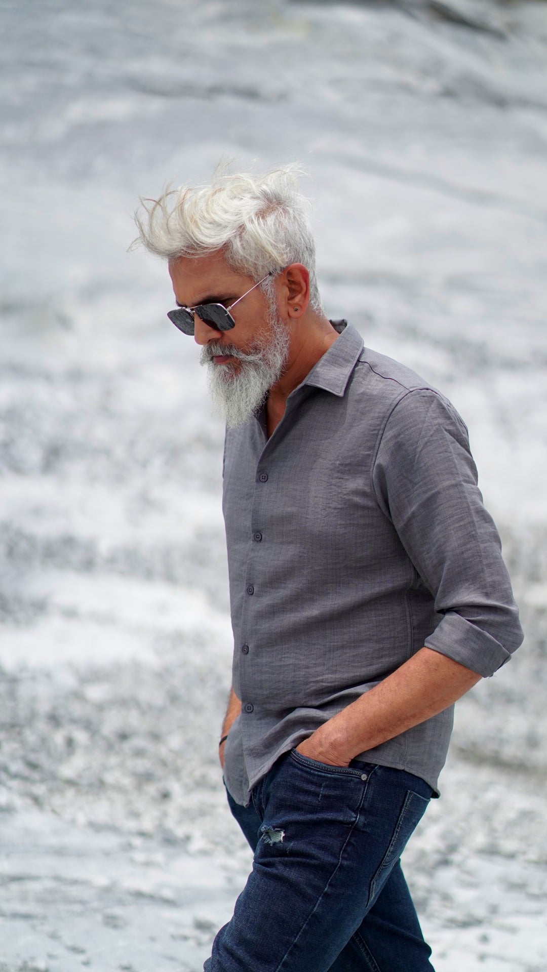 A distinguished older male model with a gray beard and stylishly combed gray hair is wearing a charcoal grey, full-sleeve shirt from Mod Maven. He pairs the shirt with dark trousers and accessorizes with black sunglasses and a sleek black wristwatch, standing confidently against a textured, natural background. The overall look exudes modern sophistication and timeless style.