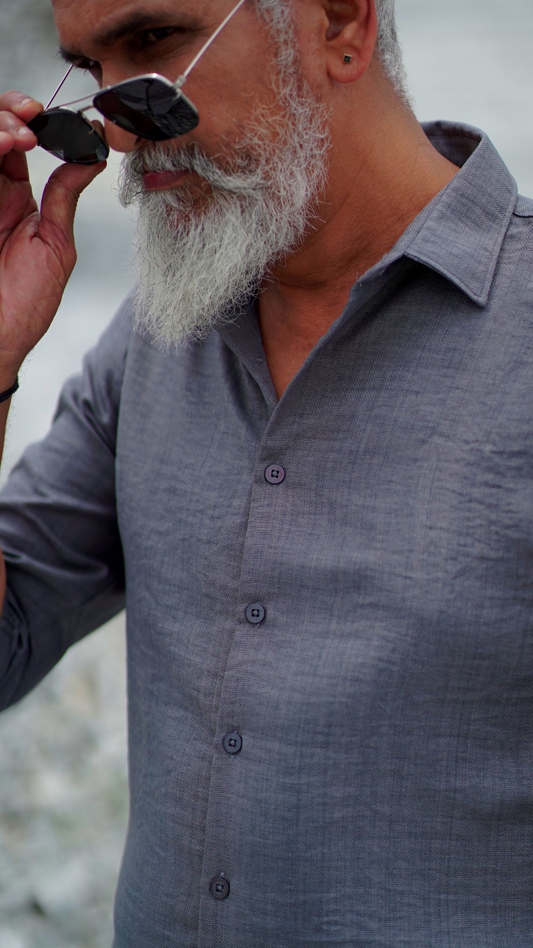 Close up shot of a distinguished older male model with a gray beard and stylishly combed gray hair is wearing a charcoal grey, full-sleeve shirt from Mod Maven. He pairs the shirt with dark trousers and accessorizes with black sunglasses and a sleek black wristwatch, standing confidently against a textured, natural background. The overall look exudes modern sophistication and timeless style.