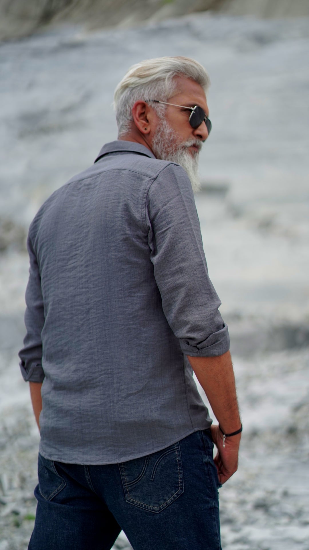 Back view of a distinguished older male model with a gray beard and stylishly combed gray hair is wearing a charcoal grey, full-sleeve shirt from Mod Maven. He pairs the shirt with dark trousers and accessorizes with black sunglasses and a sleek black wristwatch, standing confidently against a textured, natural background. The overall look exudes modern sophistication and timeless style.