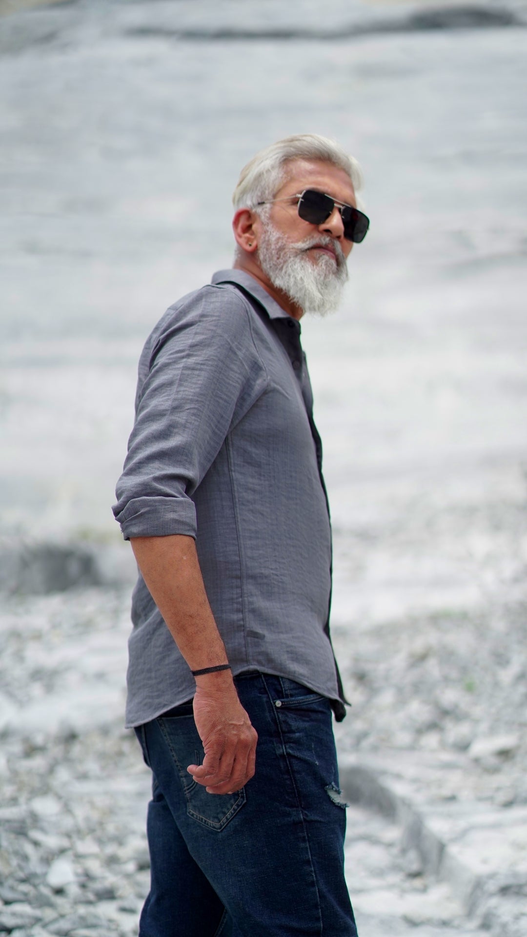 Side view of a distinguished older male model with a gray beard and stylishly combed gray hair is wearing a charcoal grey, full-sleeve shirt from Mod Maven. He pairs the shirt with dark trousers and accessorizes with black sunglasses and a sleek black wristwatch, standing confidently against a textured, natural background. The overall look exudes modern sophistication and timeless style.