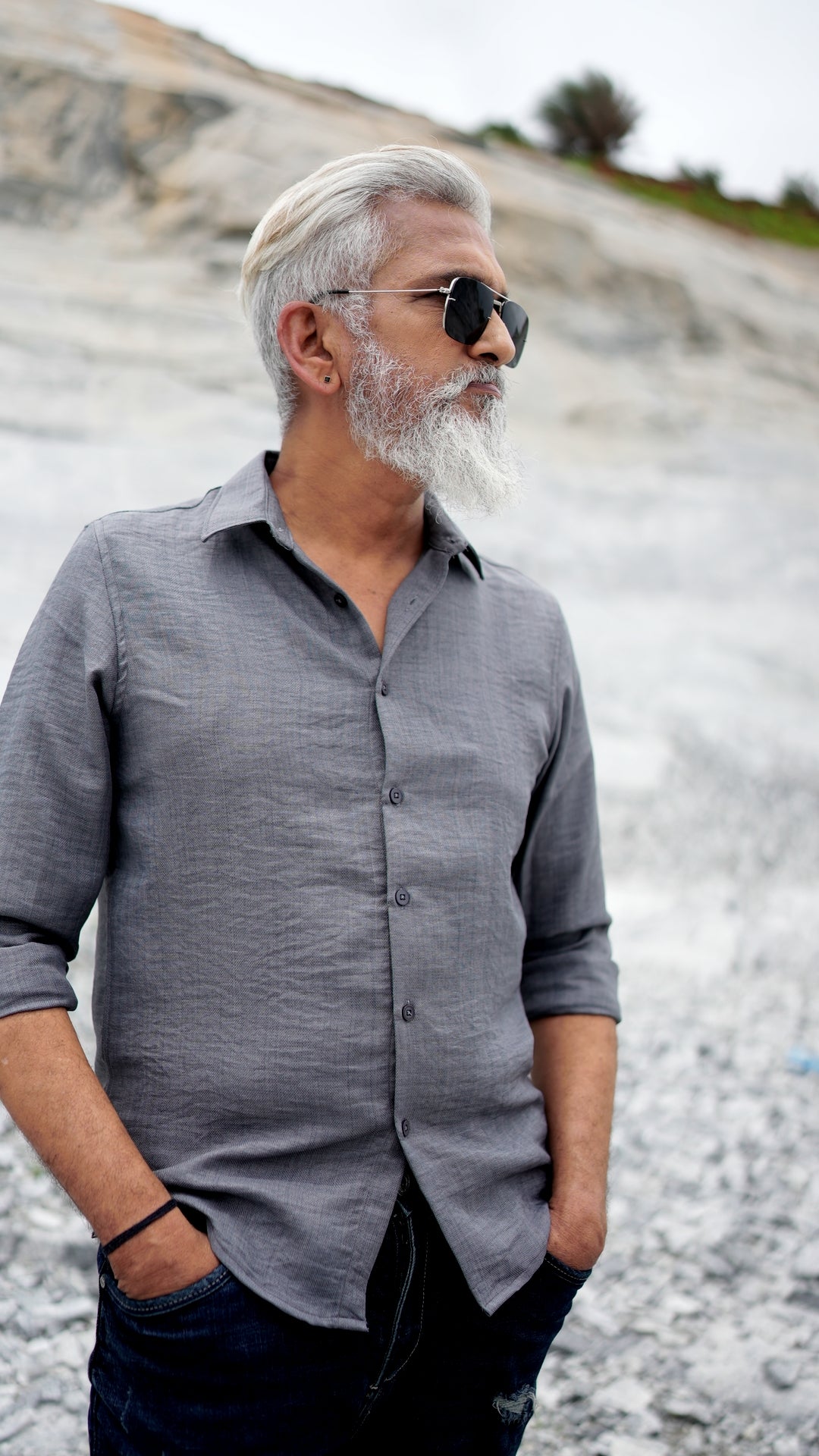 A distinguished older male model with a gray beard and stylishly combed gray hair is wearing a charcoal grey, full-sleeve shirt from Mod Maven. He pairs the shirt with dark trousers and accessorizes with black sunglasses and a sleek black wristwatch, standing confidently against a textured, natural background. The overall look exudes modern sophistication and timeless style.