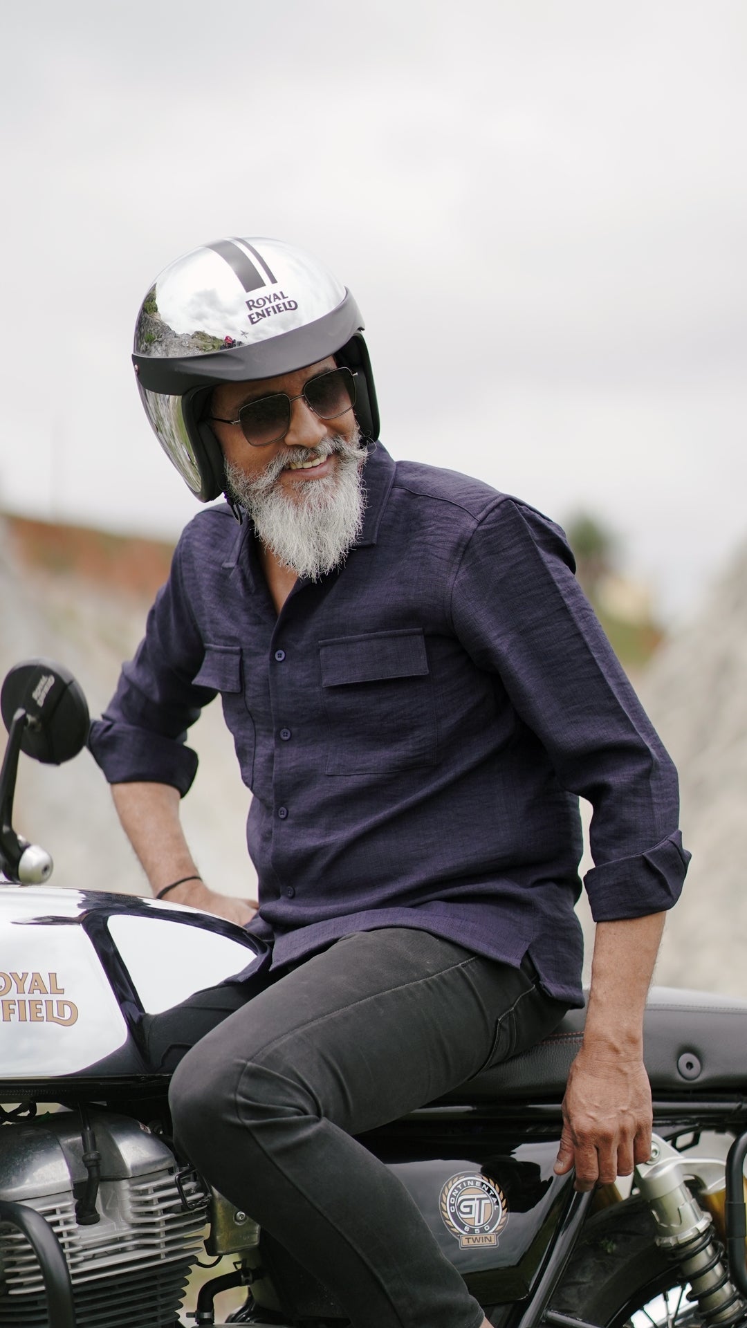 An older male model with a gray beard is wearing a navy blue, double-pocket, full-sleeve shirt from Mod Maven. He pairs the shirt with dark trousers and accessorizes with black sunglasses, sitting on a Royal Enfield Continental Gt  wearing a metallic finish, confidently against a textured, natural background. The overall look exudes modern sophistication and timeless style.