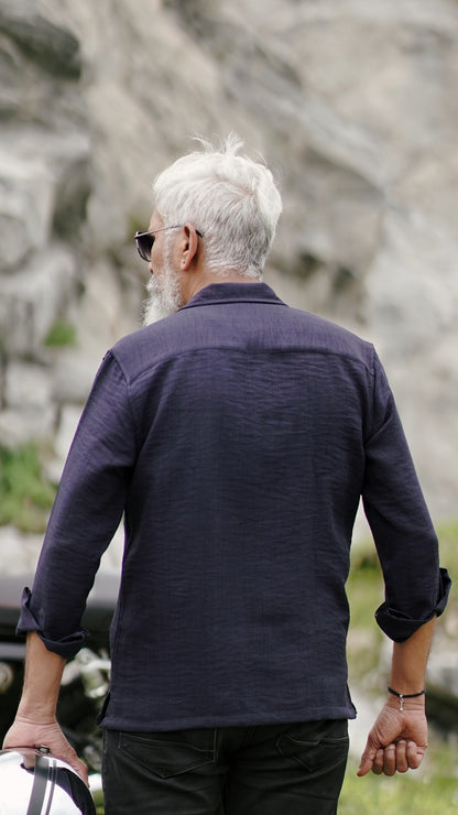 Back view of a stylish older male model with a neatly combed gray hair is showcasing an navy blue, double-pocket, full-sleeve shirt from Mod Maven. Set against a rocky backdrop conveying a sense of adventure and elegance. His outfit is completed with black trousers, black shoes,  emphasizing his effortlessly cool and sophisticated style.