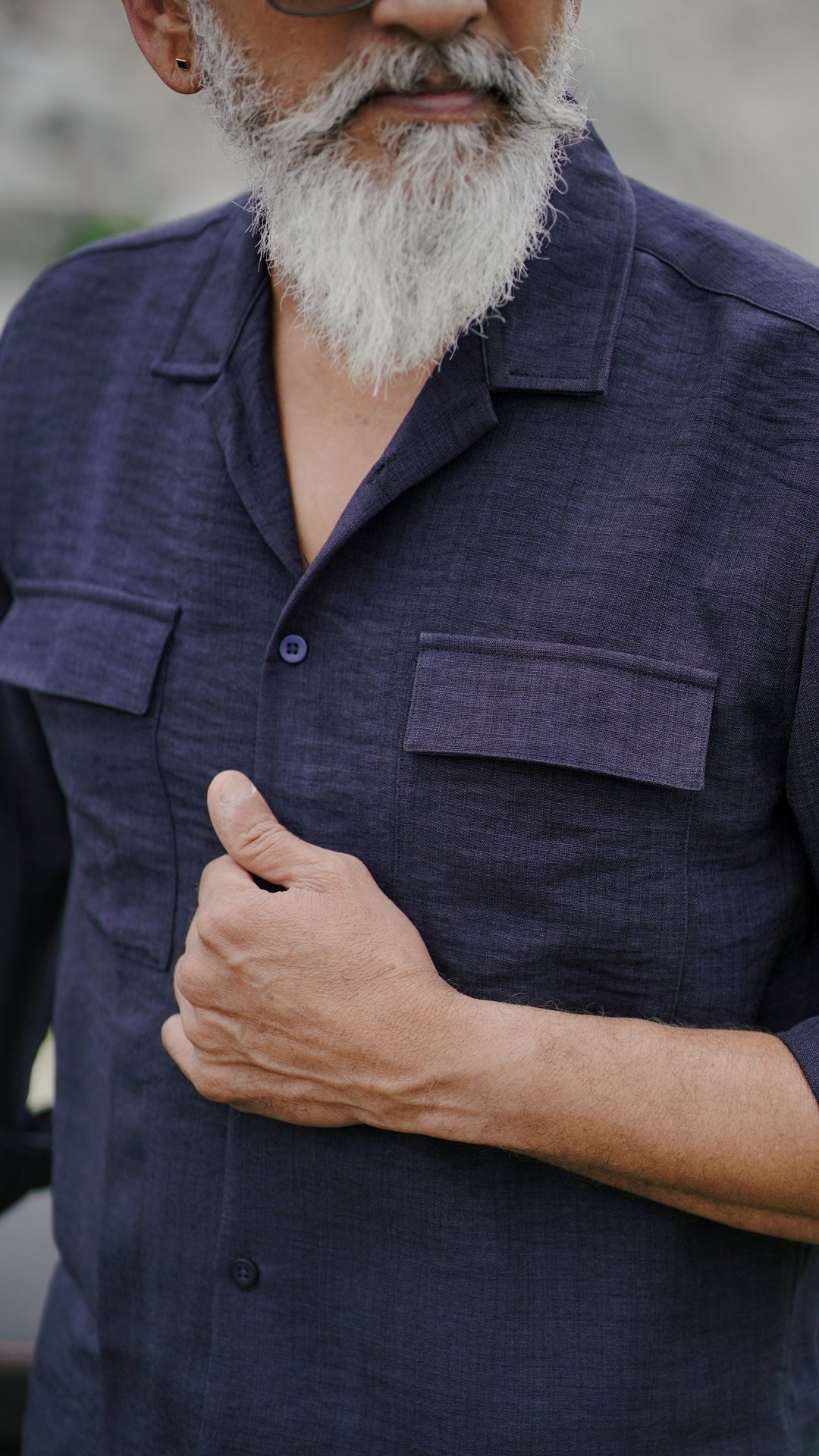 A distinguished older male model with a gray beard and stylishly combed gray hair is wearing an navy blue, double-pocket, full-sleeve shirt from Mod Maven, standing confidently against a textured, natural background. The picture focuses on the collar and pocket detail