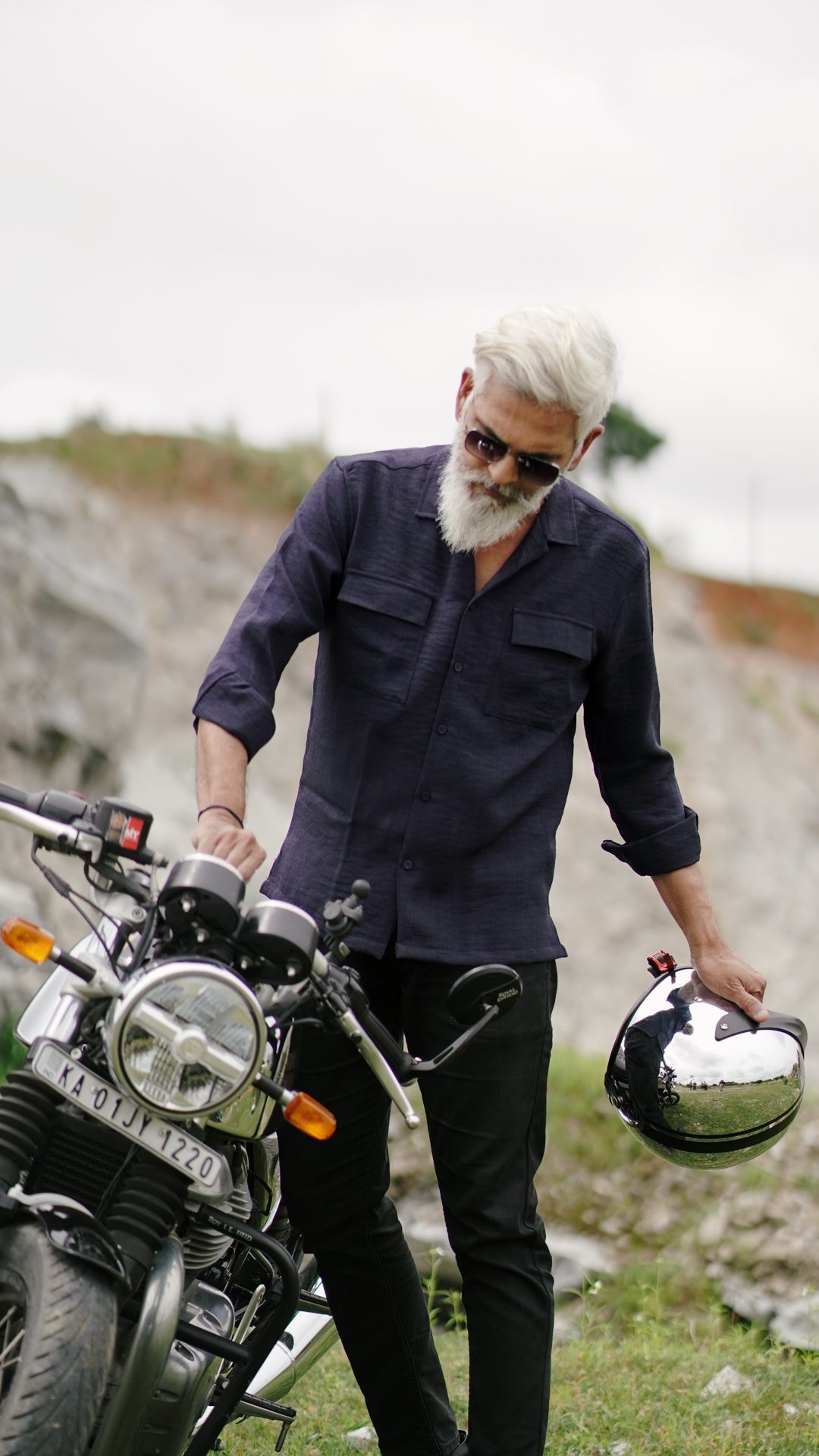 An older male model with a gray beard is wearing a navy blue, double-pocket, full-sleeve shirt from Mod Maven. He pairs the shirt with dark trousers and accessorizes with black sunglasses, switching on a Royal Enfield Continental GT , confidently against a textured, natural background. The overall look exudes modern sophistication and timeless style.