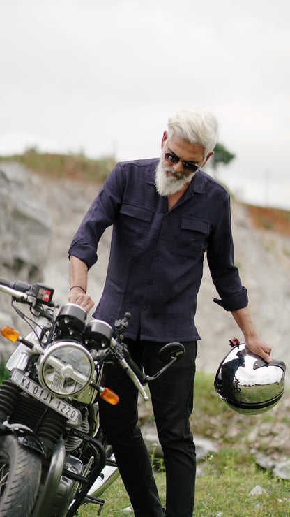 An older male model with a gray beard is wearing a navy blue, double-pocket, full-sleeve shirt from Mod Maven. He pairs the shirt with dark trousers and accessorizes with black sunglasses, switching on a Royal Enfield Continental GT , confidently against a textured, natural background. The overall look exudes modern sophistication and timeless style.