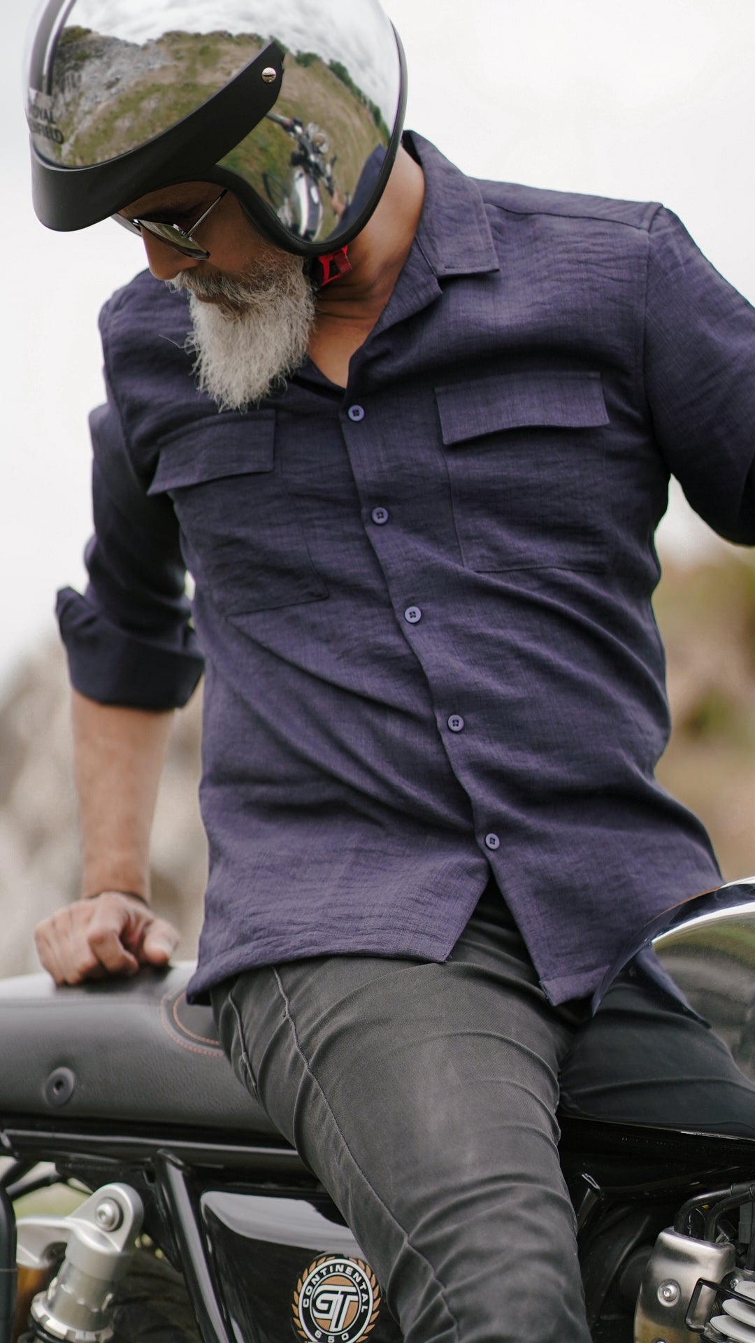 An older male model with a gray beard is wearing a navy blue, double-pocket, full-sleeve shirt from Mod Maven. He pairs the shirt with dark trousers and accessorizes with black sunglasses, sitting on a Royal Enfield Continental Gt  wearing a metallic finish helmet, confidently against a textured, natural background. The overall look exudes modern sophistication and timeless style.