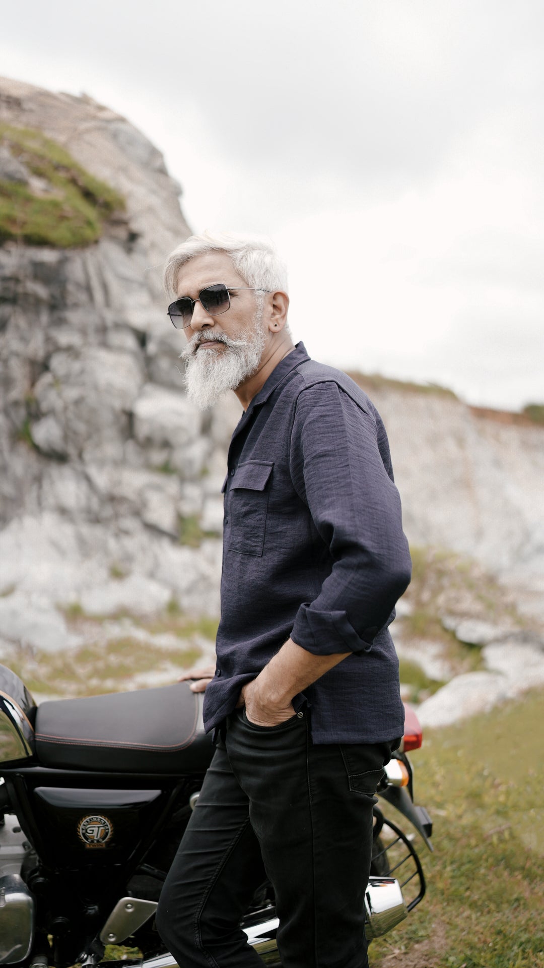 An older male model with a gray beard is wearing a navy blue, double-pocket, full-sleeve shirt from Mod Maven. He pairs the shirt with dark trousers and accessorizes with black sunglasses, standing near a Royal Enfield Continental Gt , confidently against a textured, natural background. The overall look exudes modern sophistication and timeless style.