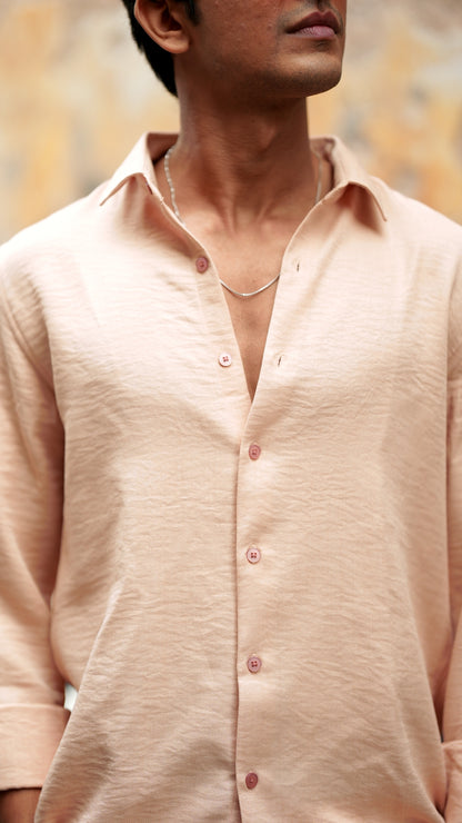 Close-up view of a male model wearing a light peach, slim-fit, full-sleeve shirt from Mod Maven. The shirt is buttoned down with matching peach buttons, showing a sleek, minimalistic design. He is accessorized with a thin silver chain, adding a touch of elegance to the outfit. The background features a subtle, neutral wall that complements the soft tones of the shirt