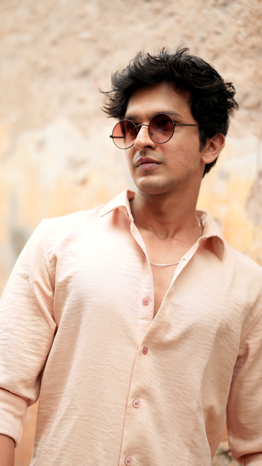 A male model wearing a light peach, slim-fit, full-sleeve shirt from Mod Maven. He has short, tousled black hair, and wears round, tinted sunglasses. The shirt has a soft texture with matching peach buttons. He is standing in front of a neutral-toned wall, exuding a casual yet refined look.