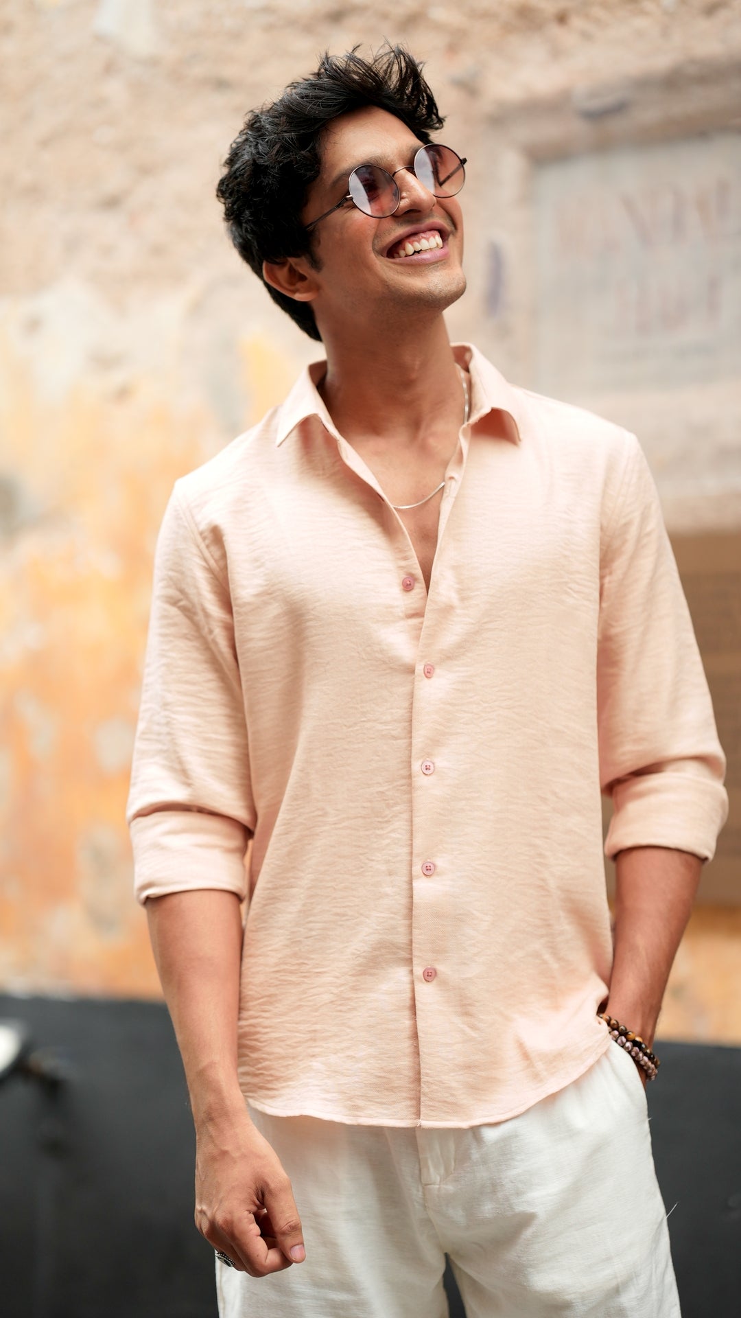 A male model wearing a light peach, slim-fit, full-sleeve shirt from Mod Maven. He has short, tousled black hair, shirt paired with linen off white trousers and wears round, tinted sunglasses. The shirt has a soft texture with matching peach buttons. He is standing in front of a neutral-toned wall, exuding a casual yet refined look.