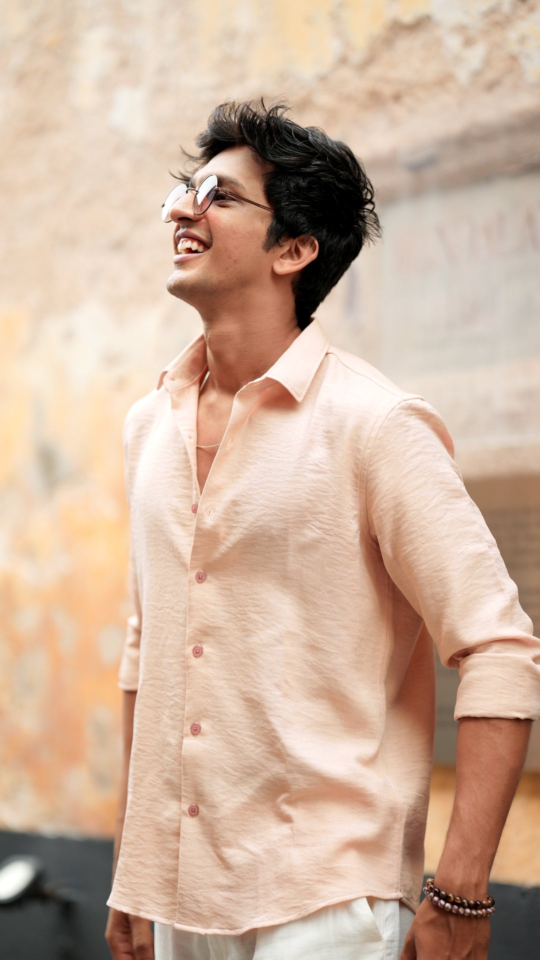 Side look of a male model wearing a light peach, slim-fit, full-sleeve shirt from Mod Maven. He has short, tousled black hair, and wears round, tinted sunglasses. The shirt has a soft texture with matching peach buttons. He is standing in front of a neutral-toned wall, exuding a casual yet refined look.