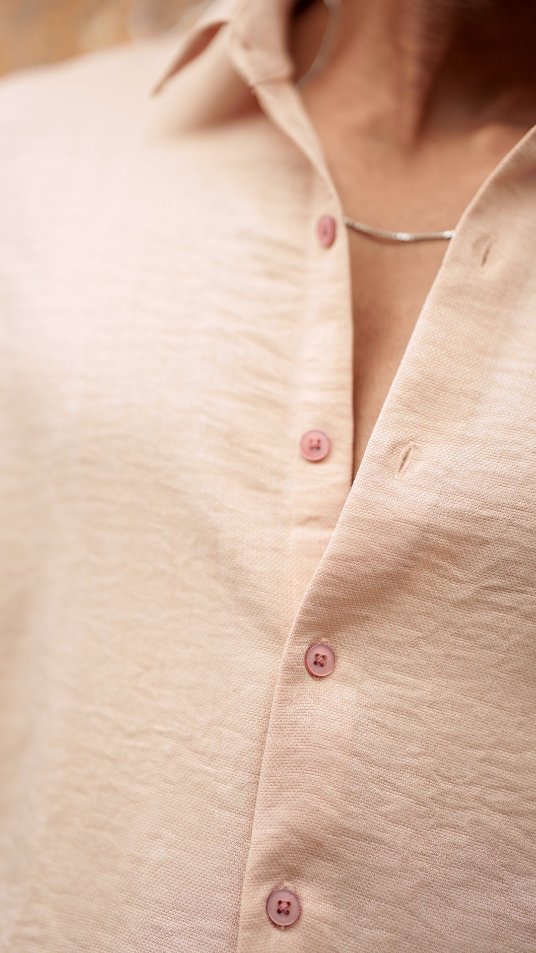 Close-up view of a male model wearing a light peach, slim-fit, full-sleeve shirt from Mod Maven. The shirt is buttoned down with matching peach buttons, showing a sleek, minimalistic design. He is accessorized with a thin silver chain, adding a touch of elegance to the outfit. The background features a subtle, neutral wall that complements the soft tones of the shirt