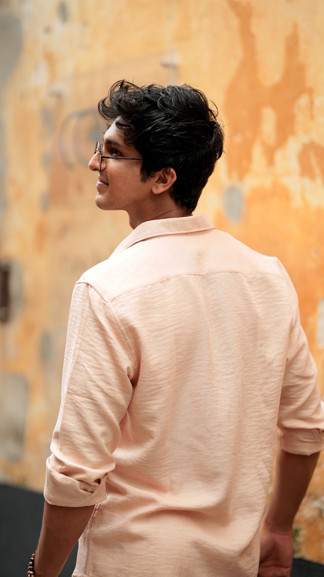 Back view of a male model wearing a light peach, slim-fit, full-sleeve shirt from Mod Maven. He has short, tousled black hair, and wears round, tinted sunglasses. The shirt has a soft texture with matching peach buttons. He is standing in front of a neutral-toned wall, exuding a casual yet refined look.