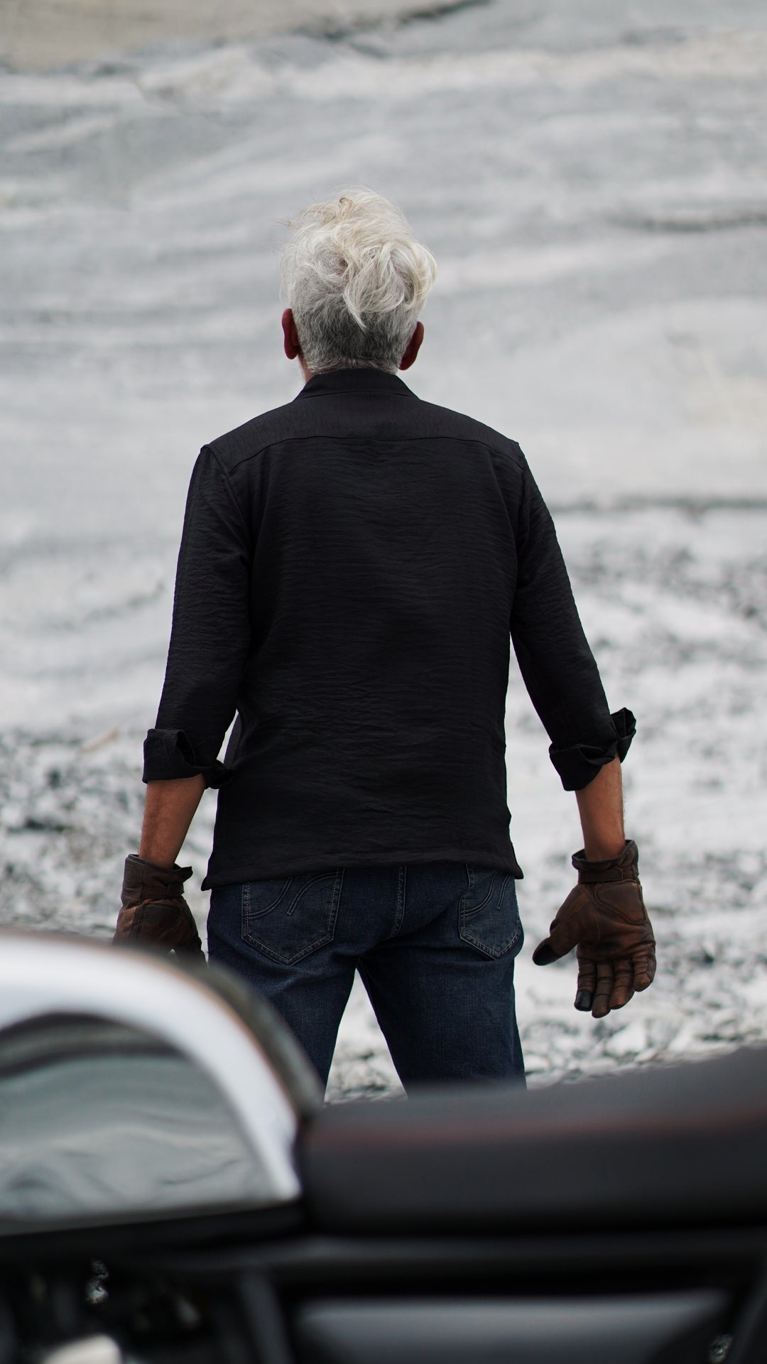 Back view of a stylish older male model with a neatly combed gray hair is showcasing an black, double-pocket, full-sleeve shirt from Mod Maven. Set against a rocky backdrop conveying a sense of adventure and elegance. His outfit is completed with black trousers, black shoes,  emphasizing his effortlessly cool and sophisticated style.