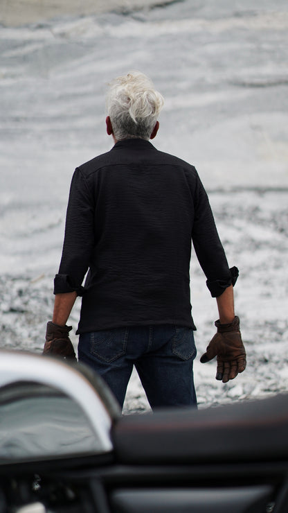 Back view of a stylish older male model with a neatly combed gray hair is showcasing an black, double-pocket, full-sleeve shirt from Mod Maven. Set against a rocky backdrop conveying a sense of adventure and elegance. His outfit is completed with black trousers, black shoes,  emphasizing his effortlessly cool and sophisticated style.