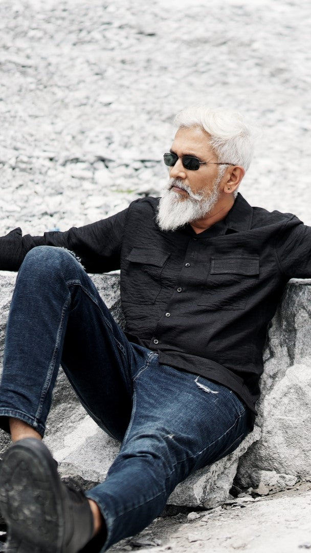 An older male model with a gray beard is wearing a black, double-pocket, full-sleeve shirt from Mod Maven. He pairs the shirt with dark trousers and accessorizes with black sunglasses, sitting on a rock  confidently against a textured, natural background. The overall look exudes modern sophistication and timeless style.