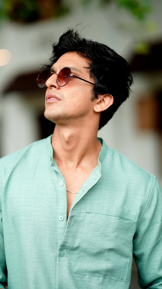 A male model wearing a pastel green, slim-fit, full-sleeve shirt from Mod Maven. He has short, tousled black hair, and wears round, tinted sunglasses. The shirt has a soft texture with matching buttons. He is standing in front of a cafe wall, exuding a casual yet refined look.