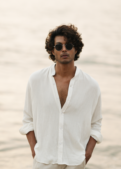 Mod Maven's white crushed cotton full sleeve shirt exudes casual elegance, paired with off-white trousers for a comfortable fit. The model stands against a serene beach backdrop, wearing stylish shades, showcasing the brand's effortlessly chic and laid-back aesthetic.