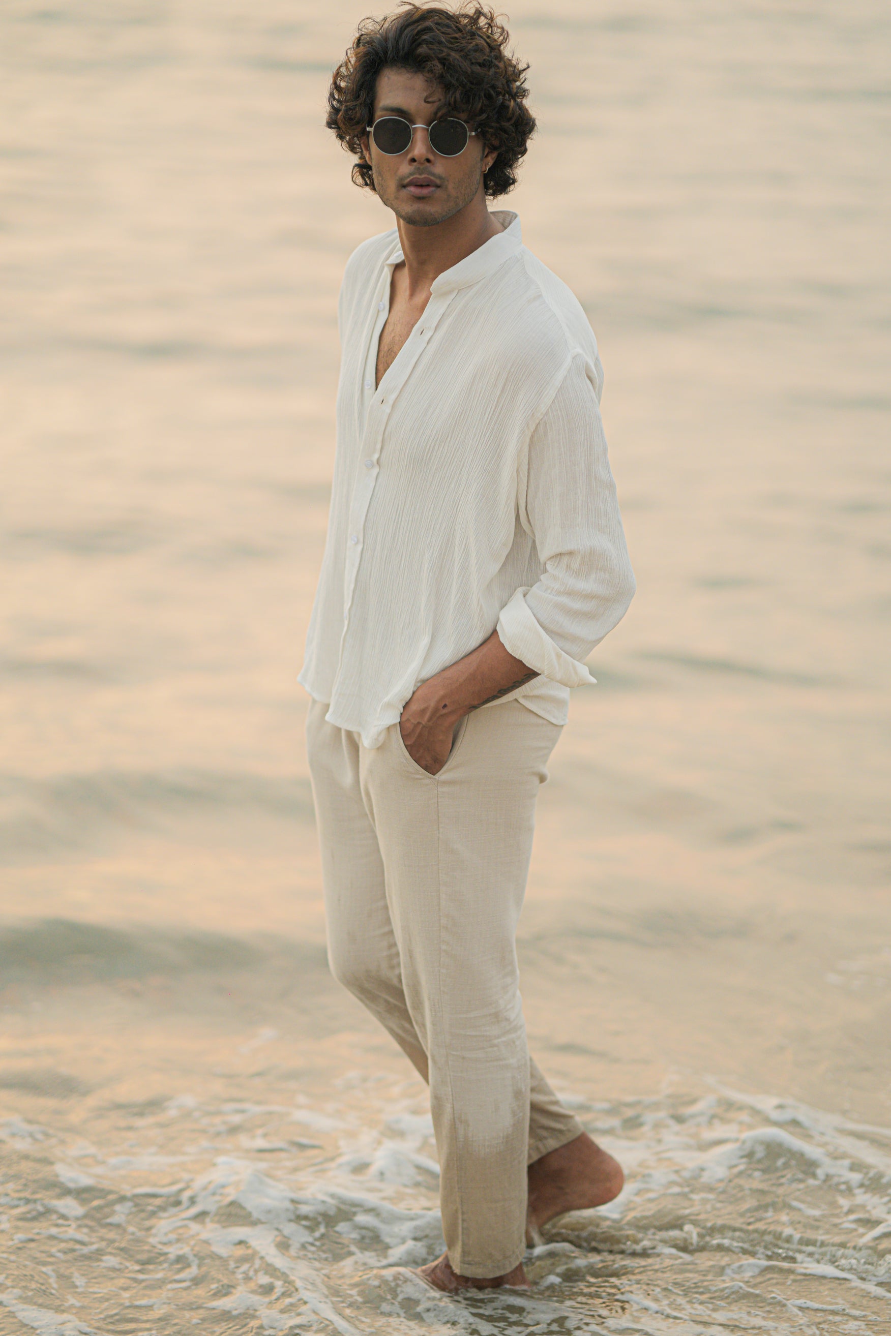 Mod Maven's white crushed cotton full sleeve shirt exudes casual elegance, paired with off-white trousers for a comfortable fit. The model stands against a serene beach backdrop, wearing stylish shades, showcasing the brand's effortlessly chic and laid-back aesthetic.