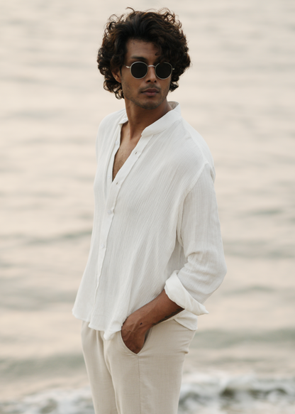 Mod Maven's white crushed cotton full sleeve shirt exudes casual elegance, paired with off-white trousers for a comfortable fit. The model stands against a serene beach backdrop, wearing stylish shades, showcasing the brand's effortlessly chic and laid-back aesthetic.