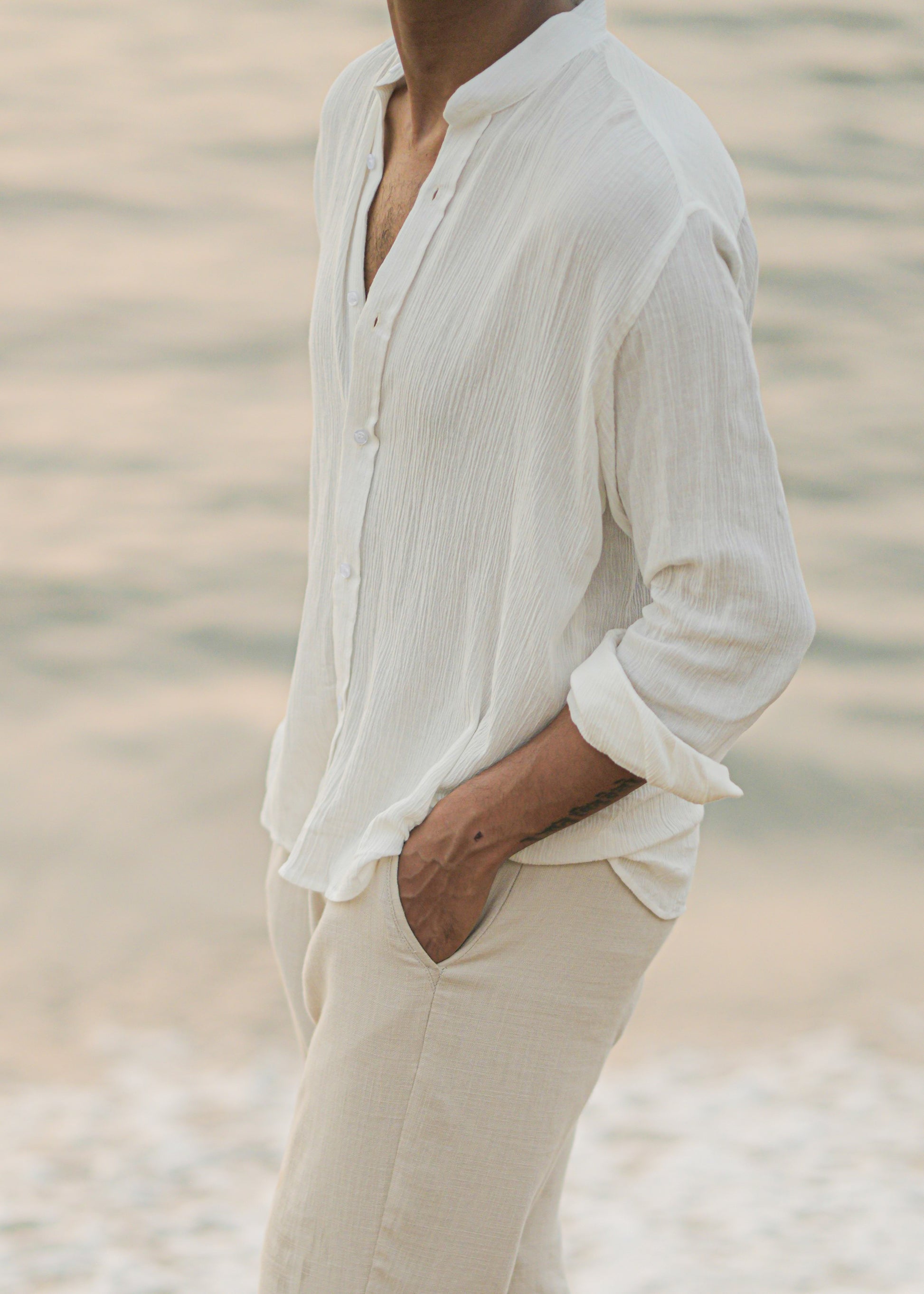 Mod Maven's white crushed cotton full sleeve shirt exudes casual elegance, paired with off-white trousers for a comfortable fit. Image shows a close up shot of model in the crushed cotton shirt, standing against a serene beach backdrop, showcasing the brand's effortlessly chic and laid-back aesthetic.